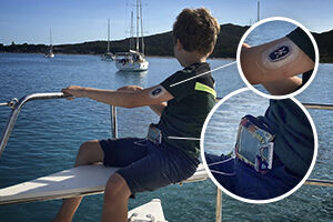 Ein Kind sitzt unter strahlend blauem Himmel beim Segeln über einen See an der Reling eines Bootes. Am linken Arm des Kindes sieht man einen Sensor, um den Bauch ist ein Messgerät geschnallt.