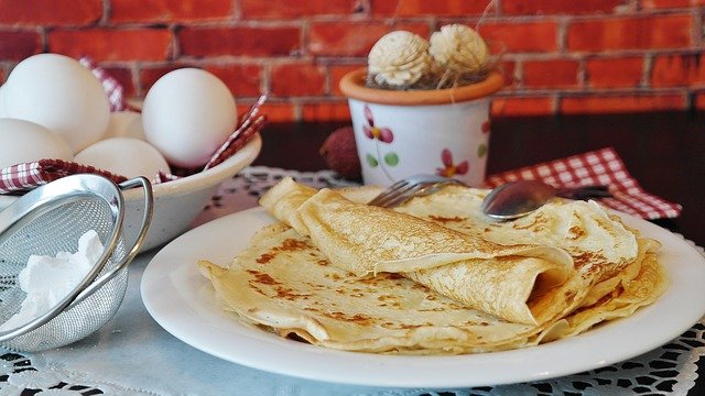 Pfannkuchen auf einem Teller gestapelt, der oberste Pfannkuchen ist zusammengerollt. Im Hintergrund steht ein Schälchen mit gekochten Eiern, eine unauffällige Blumendeko und neben dem Teller ein Sieb mit Puderzucker zum Bestäuben der Pfannkuchen.