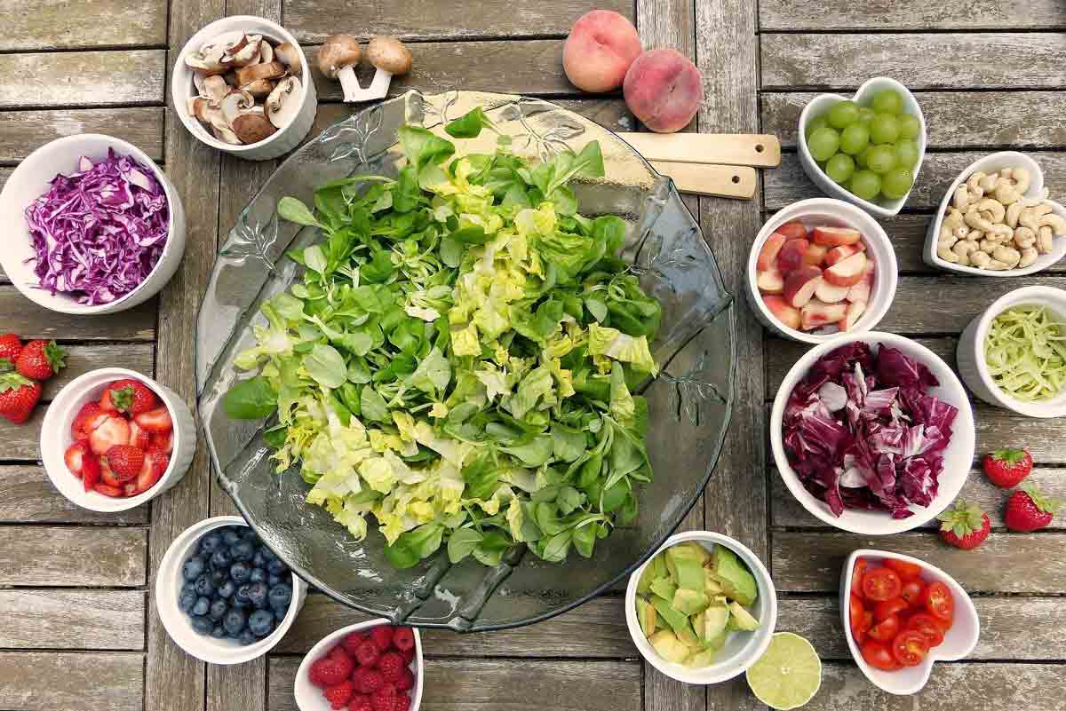 Um eine große Salatschüssel mit gemischtem grünen Salat stehen viele kleine Schälchen mit unterscheidnlichen Inhalten. Darunter Blaubeeren oder Heidelbeeren, Himbeeren, Erdbeere, Nüsse und Obst wie Pfirsiche. Außerdem noch Avocado, Tomaten und Pilze.